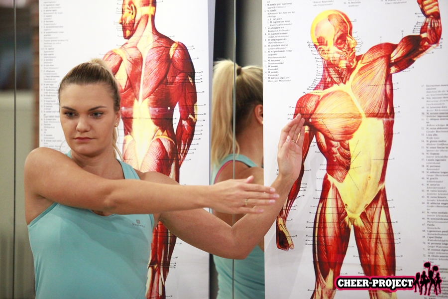 Kobieta wskazuje na swoją rękę, w tle plakat rozkładu mięśni człowieka